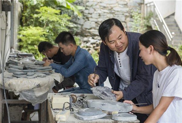 A She inkstone artisan demonstrating stone carving to an apprentice at a workbench in Wuyuan
