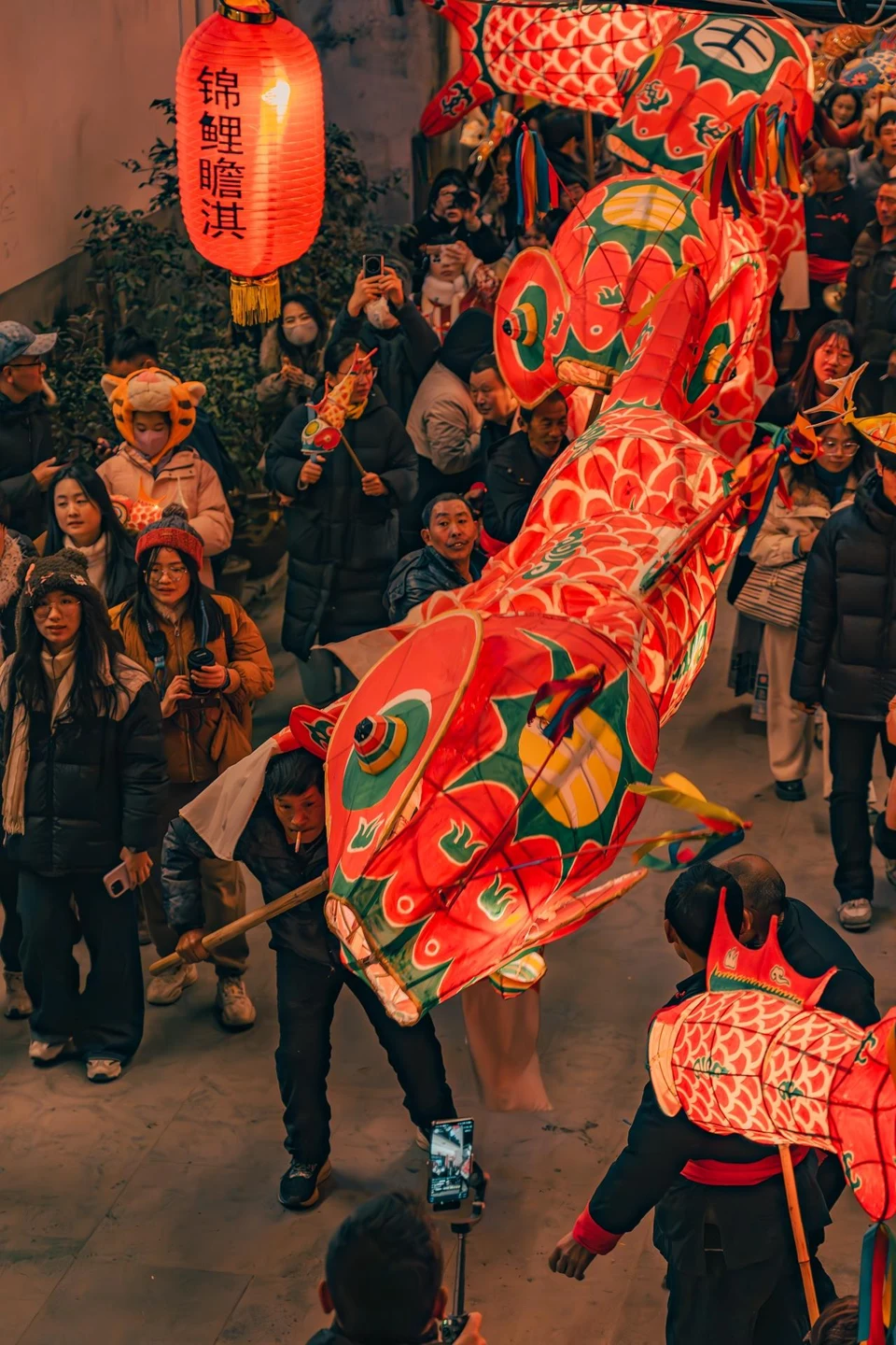 People carrying illuminated fish lanterns through a crowded lane during a Zhanqi village festival