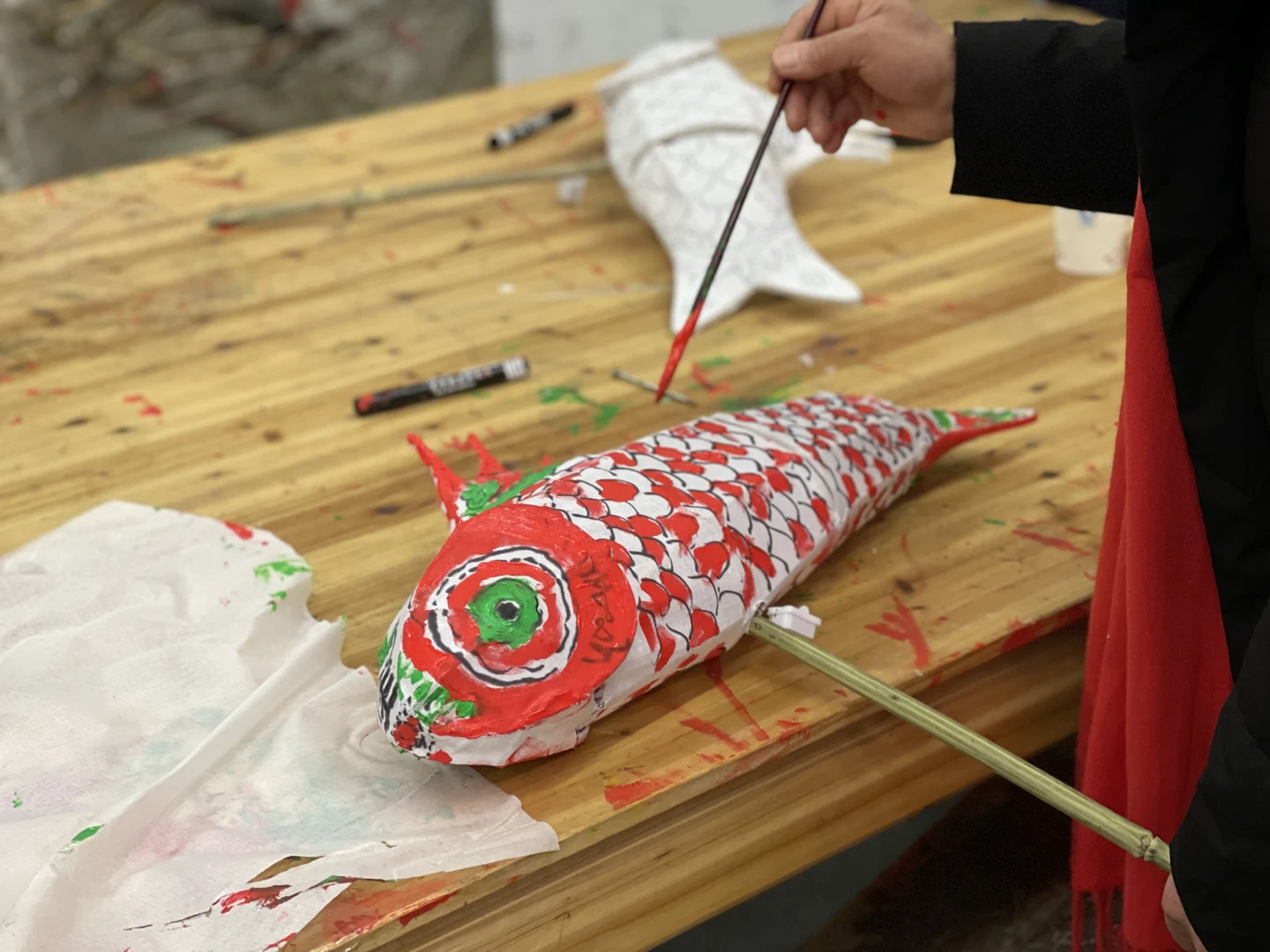 A hand painting red scales onto a paper fish lantern at a wooden workshop table