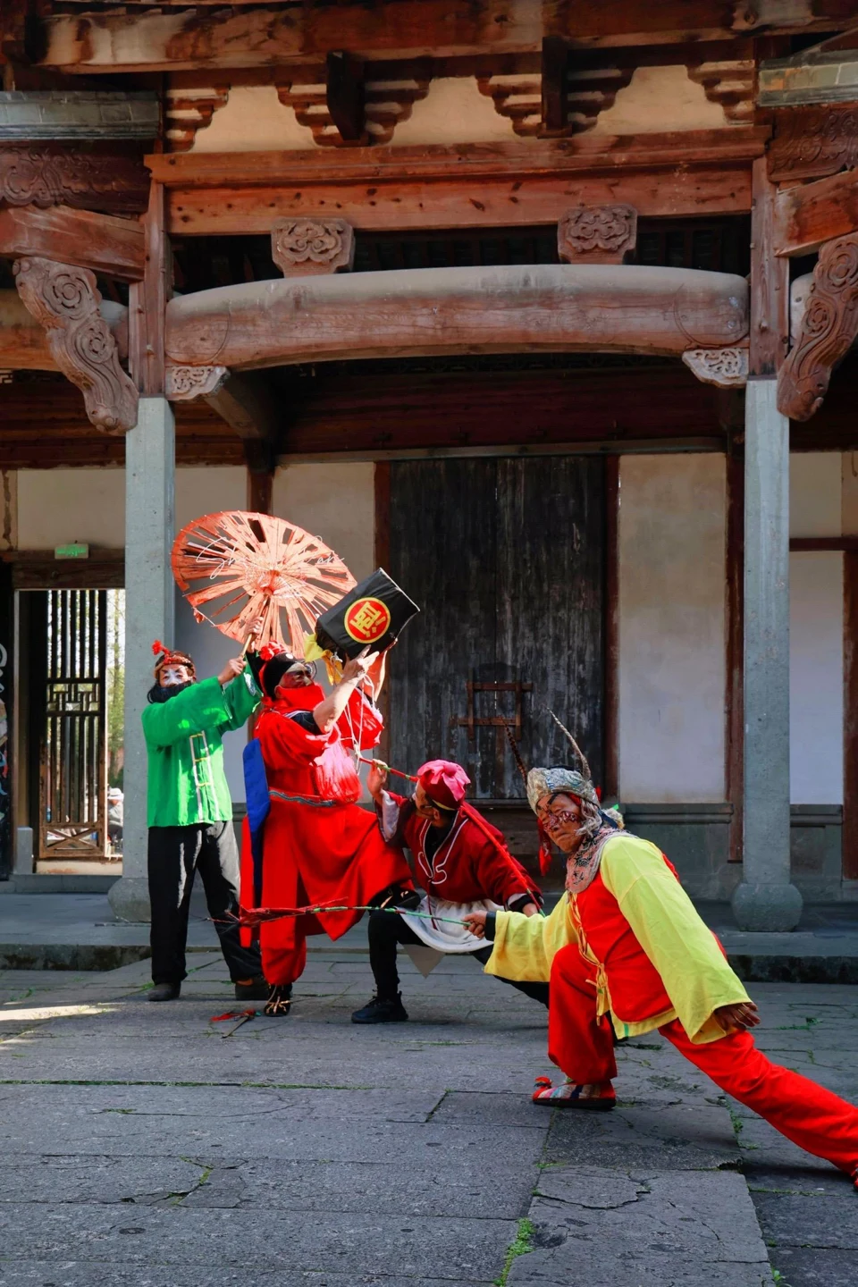 Zhongkui performers in red, green and yellow costumes moving in front of a carved timber gateway